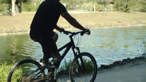 Side-view-of-man-with-artificial-leg-cycling-along-river-in-park